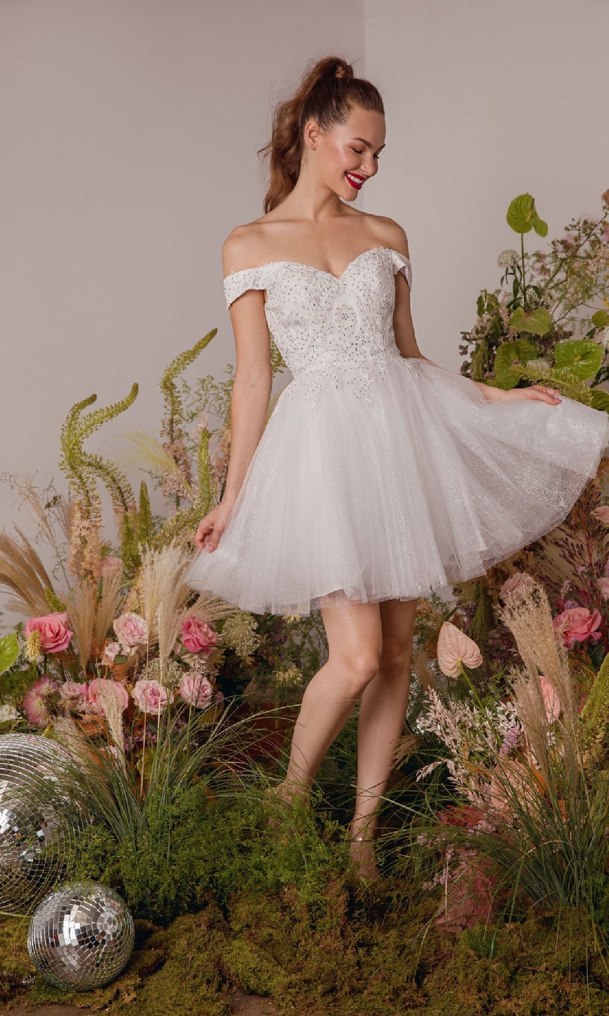 A woman wearing the Off-the-Shoulder Short Hoco Dress: Eureka 9833 smiles among flowers, greenery, and disco balls.
